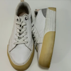 Vita sneakers med beige sula - Säljer ett par vita sneakers med beige sula och grå detaljer. De har en snygg snörning och en liten rosa detalj vid hälen. Perfekta för både vardag och fest! Skorna är i bra skick och redo för nya äventyr.