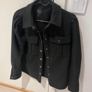 Svart jacka från Only - Säljer en snygg svart jacka från Only i bra skick. Den har långa ärmar och knappar framtill. Perfekt för höst och vår, med en stilren design som passar till det mesta. Jackan har två praktiska bröstfickor med knappar. Använd 2-3 gånger.