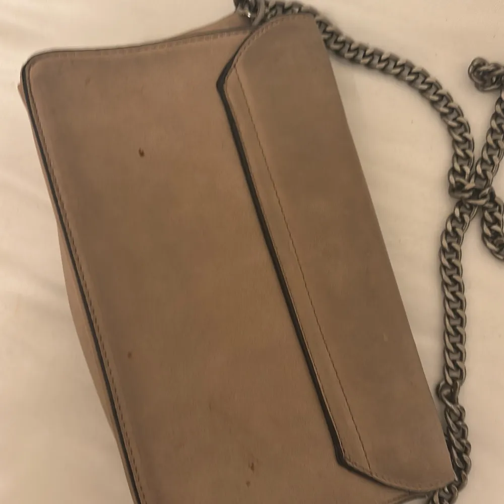 Säljer en snygg beige axelväska med en cool kedjerem. Väskan har en stilren design med ett dekorativt spänne på framsidan. Perfekt för både vardag och fest! 👜men är lite smutsig på baksidan 🤪💞. Väskor.