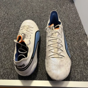 Vita fotbollsskor med blå detaljer - Säljer ett par vita fotbollsskor från Puma i bra skick. Skorna har blå och svarta detaljer samt en cool hexagonmönstrad yta. Perfekta för fotbollsträning eller match. Några tecken på användning men fortfarande i bra skick. Skostorlek EU 43. Där den har låssnat går det att limma ihop