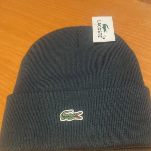 Mörkblå stickad mössa från Lacoste - Snygg och stilren mörkblå stickad mössa från Lacoste. Perfekt för kyliga dagar med sin mjuka och bekväma passform. Den har Lacostes ikoniska krokodillogga broderad framtill. Ett måste för vintern!
