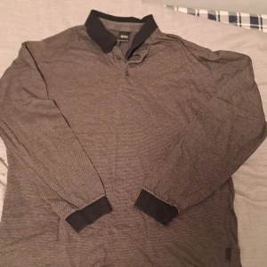 Grå pikétröja från Hugo Boss - Säljer en stilren grå pikétröja från Hugo Boss i merceriserad bomull. Tröjan har långa ärmar och en klassisk krage med knappar. Perfekt för både vardag och lite finare tillfällen. Passar bra till jeans eller chinos! Den är I ny skick bara använd 5 gånger och är i xl barn 