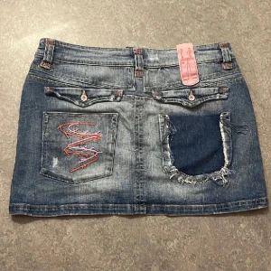 Jeanskjol med slitna detaljer - Säljer en cool jeanskjol med slitna detaljer och broderi på bakfickan. Kjolen har en klassisk blå jeansfärg och är perfekt för en avslappnad stil. Passar perfekt till sommaren eller våren! bra skick, köpt på plick men säljer igen då den är för stor för mig. Bilderna är från förra ägaren. midja: cirka 82cm höft: cirka 96cm längd: 35cm