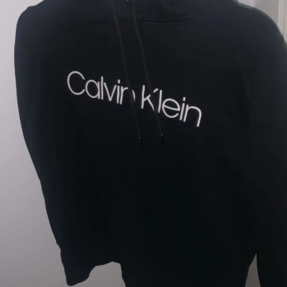 Säljer en stilren svart hoodie från Calvin Klein. Den har en klassisk passform med justerbar huva och det ikoniska Calvin Klein-logotypen tryckt på framsidan. Perfekt för en avslappnad look eller att mysa i hemma. Passar till alla säsonger!. Neuletakit & Villapaidat.