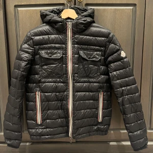 Svart dunjacka från Moncler - Säljer en snygg svart dunjacka från Moncler i mycket bra skick. ”Moncler douvert”. Jackan har en quiltad design med dragkedja och huva, samt praktiska fickor med knappar. Perfekt för kyliga dagar och ger en stilren look. Storlek 2