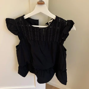 Svart blus med volangdetaljer - Säljer en söt svart blus med volangdetaljer på ärmarna och nederkanten. Blusen har en fin struktur upptill och är perfekt för både vardag och fest. Den är i bra skick och passar till många olika outfits.
