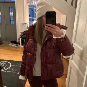 Vinröd pufferjacka - Säljer en snygg vinröd pufferjacka i glansigt material. Jackan har en dragkedja framtill och är perfekt för kalla dagar. Den är långärmad och har en mysig, vadderad känsla. Perfekt för höst och vinter!