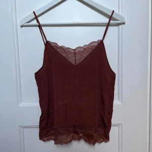 Brun linne med spetsdetaljer - Snygg brun linne med tunna axelband och v-ringad spetskant både fram och bak. Perfekt för sommardagar eller som ett lager under en kavaj. Materialet känns mjukt och luftigt, vilket gör den bekväm att bära hela dagen.