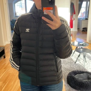 Svart dunjacka från Adidas - Säljer en snygg svart dunjacka från Adidas i storlek 38 som är använd ett fåtal gånger. Jackan har de klassiska tre vita ränderna längs ärmarna och en dragkedja framtill samt fickor med dragkedja. Perfekt för kyliga dagar med sin varma och bekväma passform. Adidas-loggan syns tydligt på bröstet och är insydd. Passar både till vardags och sportiga aktiviteter 🌟