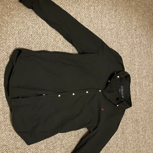 Svart skjorta från Ralph Lauren - Säljer en stilren svart skjorta från Ralph Lauren i herrstorlek M. Skjortan har långa ärmar och knappar framtill. Den är i mycket bra skick och har en liten röd broderad logga på bröstet. Perfekt för både vardag och fest!