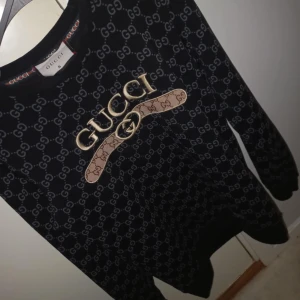 Svart tröja från Gucci - Säljer en snygg svart tröja från Gucci med deras ikoniska logotypmönster över hela tyget. Tröjan har långa ärmar och en rund halsringning. Perfekt för att ge din outfit en lyxig touch. Passar både till vardags och fest!
