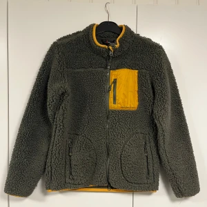 Grön piléfleece från High Mountain - Mysig mörkgrön piléfleece från High Mountain med kontrasterande oranga detaljer. Jackan har en praktisk dragkedja och en orange bröstficka med dragkedja. Perfekt för kyliga höst- och vinterdagar. Den är i mycket bra skick och redo för nya äventyr!