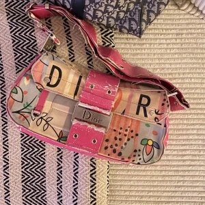 Dior väska - Säljer en unik och färgglad vintage axelväska från Dior med ett coolt mönster och text. Väskan har en bred rosa axelrem och detaljer i metall. Väskan är i acceptabelt skick med lite slitage på remmen. Välkomna med egna bud 💖