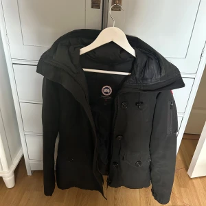 Svart jacka från Canada Goose - Säljer en svart Canada Goose jacka i mycket bra skick. Den är perfekt för kalla vinterdagar med sin varma och skyddande design. Jackan har en huva och stängs med både dragkedja och knappar. Tillverkad i polyester och bomull för extra hållbarhet. Passar perfekt för vinterns alla äventyr! 🖤