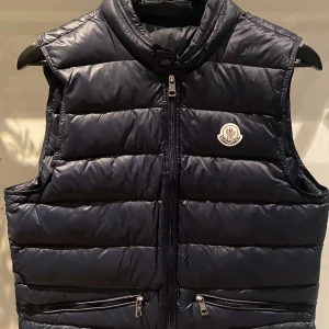 Marin blå dunväst från Moncler - Säljer en snygg svart dunväst från Moncler i mycket bra skick. Västen har en dragkedja framtill och två praktiska fickor med dragkedjor. Perfekt för höst och vår när du vill hålla dig varm och stilren. Passar både till vardags och festliga tillfällen.