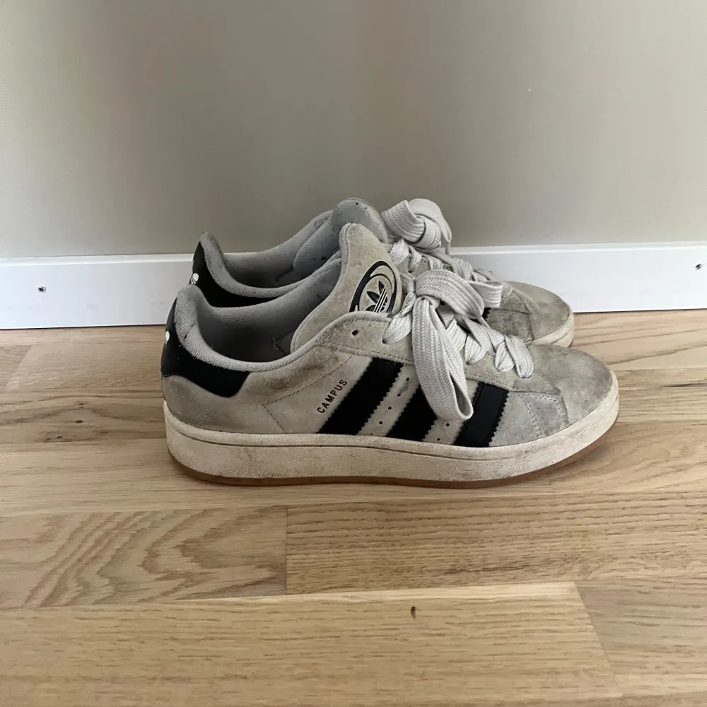 Säljer ett par klassiska Adidas Campus sneakers i beige och svart. De har en snygg retrostil med de ikoniska tre ränderna på sidan. Skorna är i bra skick men har lite slitage på sulan och ovandelen går även att skrubba lite på dom så ser dom snyggare ut. Perfekta för vardagsbruk och ger en cool touch till vilken outfit som helst. . Skor.