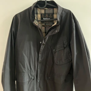 Svart Field jacket från Barbour - Säljer en snygg svart Barbour jacka i storlek L men den är liten så sitter som M. Den har en klassisk design med knappar och en stilren krage. Insidan har ett rutigt mönster som ger en extra touch. Perfekt för höst och vår!