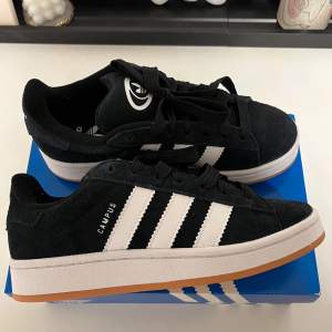 Säljer ett par snygga svarta Adidas Campus sneakers i mocka med de klassiska vita ränderna på sidan. Skorna har en vit sula med en gummifärgad undersida och kommer med både svarta och vita skosnören. Perfekta för vardagsbruk och ger en stilren look. De är i helt nytt skick och levereras i originalkartong. 