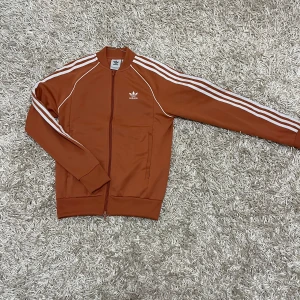  Adidas kofta - Säljer en snygg Adidas kofta i mycket bra skick. Den har de klassiska tre vita ränderna längs ärmarna och en dragkedja framtill. Perfekt för en sportig look eller vardagsstil. Koftan är långärmad och gjord i ett bekvämt syntetmaterial.