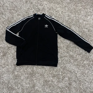 Adidas kofta - Säljer en klassisk svart Adidas jacka med de ikoniska vita ränderna längs ärmarna. Jackan har dragkedja och Adidas-loggan på bröstet. Passar till alla säsonger och ger en sportig look!