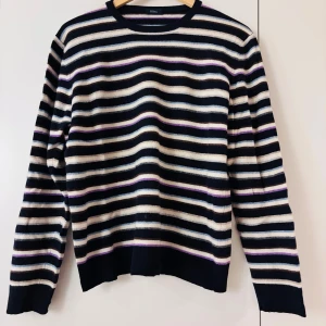 Randig ulltröja från Altea - Säljer en superfin randig tröja från Altea i 100% ull. Den har en snygg kombination av svart, beige, lila och blå ränder. Perfekt för höst och vinter med sina långa ärmar och mjuka material. Tillverkad i Italien, vilket ger den en lyxig känsla. Passar både till vardags och lite finare tillfällen!