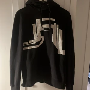 Svart hoodie från J.Lindeberg - Säljer en snygg svart hoodie från J.Lindeberg med ett coolt silvertryck på framsidan. Perfekt för både vardag och chill. Den har en klassisk passform med långa ärmar och en skön huva. Passar perfekt till jeans eller joggers! Priset kan såklart diskuteras😁