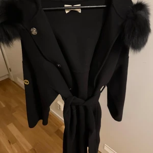 Svart kappa från Femenina of Sweden - Säljer en elegant svart kappa från Femista of Sweden. Den har en lyxig pälsdetalj på huvan och ett snyggt bälte i midjan. Kappan är långärmad och har en fin knappdetalj. Perfekt för höst och vinter! Nypris: 3900