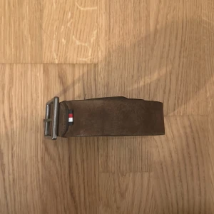 Brunt Läder Bälte  - Snyggt brunt läder bälte med ett stilrent silverfärgat spänne. Perfekt för att ge en klassisk touch till din outfit. Detalj med liten flagga i rött, vitt och blått vid spännet.