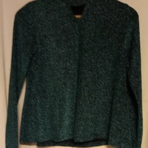 Läcker glittrig top i mörkgrön - Snygg mörkgrön glittrig top med lång ärm. Perfekt för att lägga till lite bling till din outfit. Jackan har en stilren design utan krage och är tillverkad i ett glittrande material som fångar ljuset fint.