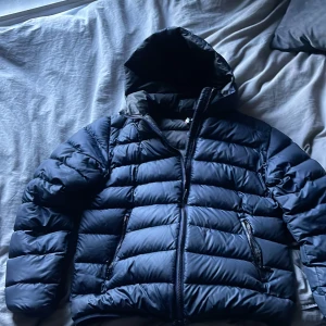 Mörkblå pufferjacka med huva - Säljer en snygg mörkblå pufferjacka från Colmar. Jackan har en praktisk huva och dragkedja framtill. Perfekt för kyliga dagar med sin quiltade design som håller värmen. Passar ungdomar med storlek 16Y eller 172 cm.