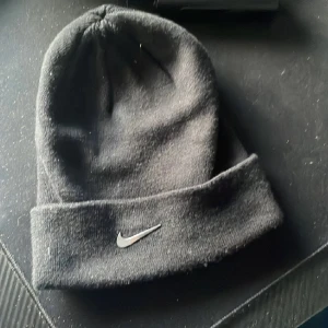 Svart mössa från Nike - Snygg svart mössa från Nike med klassisk design och uppvikt kant. Perfekt för kyliga dagar och enkel att matcha med olika outfits. Den har en diskret Nike-logga framtill.