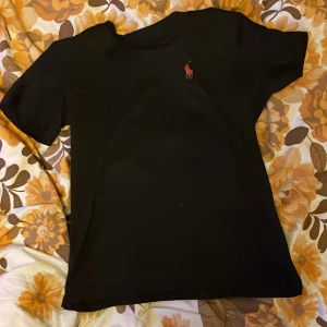 Svart t-shirt från Polo Ralph Lauren - Säljer en klassisk svart t-shirt från Polo Ralph Lauren med det ikoniska röda logotypen på bröstet. T-shirten har en normal passform och är kortärmad, perfekt för en stilren look.