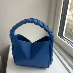 Blå axelväska med flätat handtag - Snygg blå axelväska med ett unikt flätat handtag. Väskan har en modern design med en triangulär form och är tillverkad i syntetmaterial. Perfekt för att ge din outfit en färgklick. Har magnet vi öppning vilket gör att man kan forma den på olika sätt.