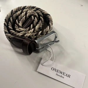 Flätat bälte från ONEWEAR - Snyggt flätat bälte från ONEWEAR i beige och brunt med en stilren silverfärgad spänne. Perfekt för att ge en extra touch till din outfit. Tillverkat i ett slitstarkt material med skinnliknande detaljer.