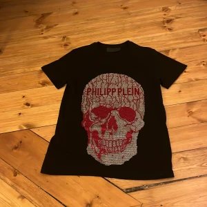 Svart t-shirt med dödskalle från Philipp Plein - Snygg svart t-shirt från Philipp Plein med en glittrig dödskalle och röd text på framsidan. Perfekt för att sticka ut med en unik stil. T-shirten har korta ärmar och en normal passform. Samt är helt oanvänd och ny. Storlek: M. Men passar även S