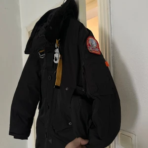 Parajumper svart med pälsdetalj - Säljer en svart parkajacka från Parajumpers med pälsdetalj på huvan. Jackan har flera fickor, dragkedja och knappar framtill samt en broderad patch på ärmen. Perfekt för kalla dagar med sin långa passform och varma material. Storlek M