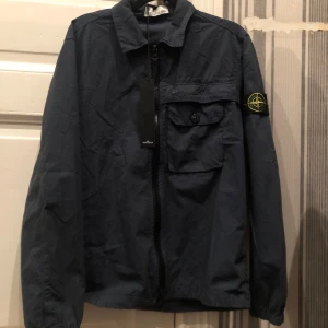Mörkblå jacka från Stone Island - Säljer en snygg mörkblå jacka från Stone Island med dragkedja och en bröstficka med knapp. Jackan har en ikonisk logga på ärmen och är perfekt för en stilren look.!det är en r3pe!
