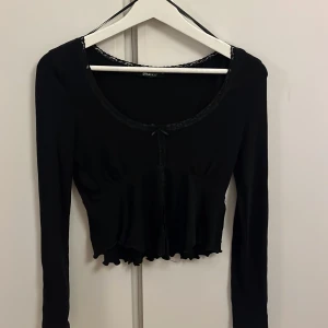 Svart långärmad topp från Gina Tricot - Säljer en svart långärmad topp från Gina Tricot. Toppen har spetsdetaljer vid halsringningen och ett snitt framtill. Den är croppad med en volangkant nedtill, vilket ger en söt och stilren look.