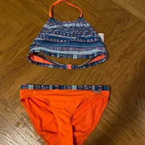 Mönstrad bikini från Firefly - Snygg bikini från Firefly med ett färgglatt mönster i blått och orange. Toppen har halterneck-design och byxorna har en mönstrade kant. Byxorna är lite fläckiga där bak, men man tänker inte på det. Perfekt för sommarens badäventyr!