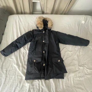 Svart parkajacka med pälsdetalj från Woolrich - Säljer en svart parkajacka från Woolrich med beige foder och pälsdetalj på huvan. Jackan har långa ärmar och flera praktiska fickor med knappar. Perfekt för kalla dagar. Den kostar egentligen 8750 men jag säljer den för 5000