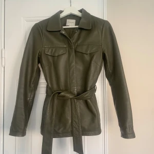 Olivgrön skinnjacka från Selected Femme - Snygg olivgrön skinnjacka från Selected Femme med knappar och bälte i midjan. Jackan har långa ärmar och två bröstfickor med lock. Perfekt för en stilren look.