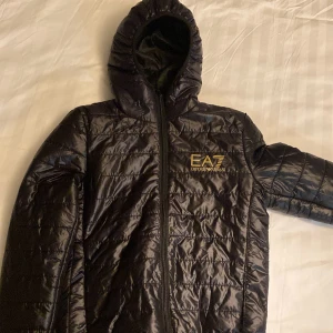Svart quiltad jacka från EA7 - Säljer en snygg svart quiltad jacka från EA7. Den har en dragkedja framtill och en huva för extra skydd mot väder. Perfekt för höst och vår, med en stilren design som passar till det mesta. Jackan är i nytt skick och redo för äventyr!