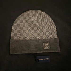 Grå rutig mössa från Louis Vuitton - Snygg grå mössa från Louis Vuitton med ett klassiskt rutigt mönster. Mössan har en uppvikt kant och ett diskret LV-märke framtill. Tillverkad i en blandning av ull och siden för en lyxig känsla.