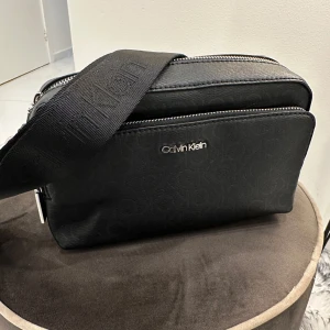 Axelremsväska från Calvin Klein  - Säljer en stilren och praktisk axelremsväska från Calvin Klein, perfekt för resor eller vardagsbruk. Väskan är både rymlig och säker, med mått som gör den smidig att bära med sig  Specifikationer: 	•	Höjd: 14 cm 	•	Längd: 22,5 cm 	•	Djup: 6 cm  Väskan är i mycket fint skick och passar lika bra till vardag som till fest. Kontakta mig vid intresse eller för fler bilder!