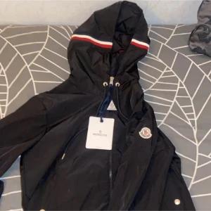 Svart jacka från Moncler - Säljer en snygg svart jacka från Moncler med huva och dragkedja. Jackan har Monclers logga på ärmen och en stilren design med röd och vit randdetalj på huvan. Perfekt för dig som vill ha något både praktiskt och trendigt. 