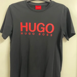 Svart t-shirt från Hugo Boss - Snygg svart t-shirt från Hugo Boss med röd logga på bröstet. Perfekt för en stilren look. Kortärmad och tillverkad i mjukt material för extra komfort. XS