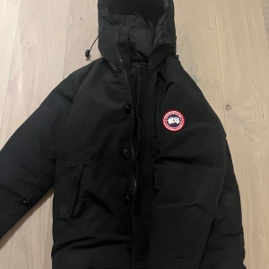 Svart dunjacka från Canada Goose - Säljer en svart dunjacka från Canada Goose med huva och dragkedja. Jackan har knappar framtill och en broderad logotyp på armen. Prutning kan tas emot