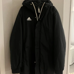 Svart jacka från Adidas - Säljer en svart parkajacka från Adidas med huva och dragkedja. Jackan har långa ärmar och Adidas-loggan på bröstet. Perfekt för utomhusaktiviteter när man vill ha nått lite sportigare och som går ner under rumpan