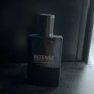 Intense Impression Eau de Toilette - Snygg och elegant flaska i svart med texten 'Intense Impression' på framsidan. Perfekt för den som vill ha en doft som gör ett starkt intryck. Flaskan har en stilren design som passar in i alla miljöer.