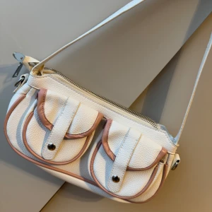 Vit axelväska med rosa detaljer - Snygg vit axelväska med rosa detaljer. Väskan har två framfickor med knappar. Har aldrig använt den. Hör av dig för mer info/bilder:)